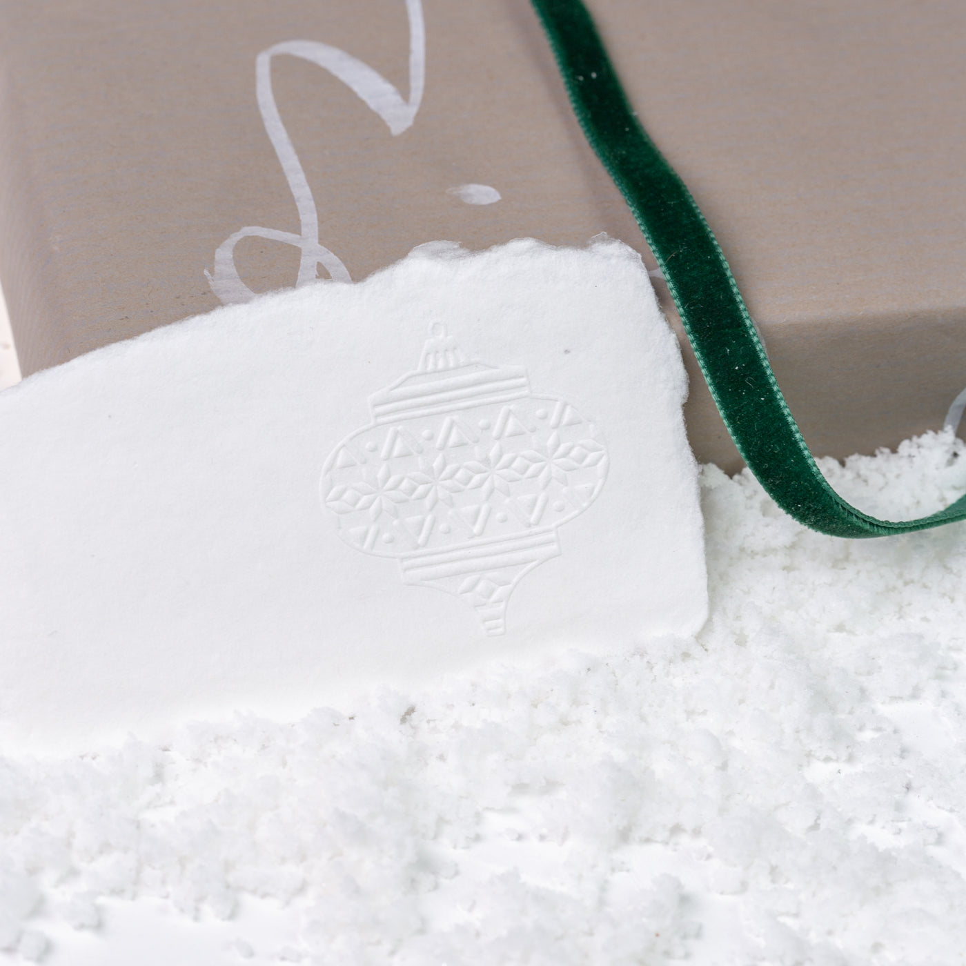 White gift tag embossed with Nordic inspired bauble tied to Christmas gift with velvet green ribbon on a bed of snow | Heirloom Seals